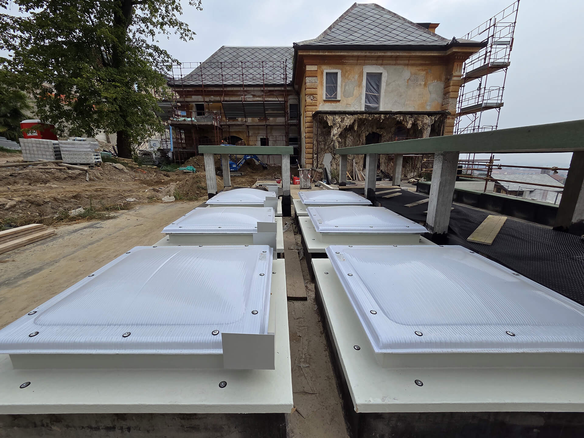 L'immagine mostra un cantiere di ristrutturazione di un edificio storico, con un focus su una serie di lucernari installati sul tetto. I lucernari sono montati su strutture metalliche, probabilmente realizzate in acciaio, e sembrano essere parte di un progetto di recupero o di aggiornamento dell'edificio. Sullo sfondo, si vede un edificio che sta subendo lavori di ristrutturazione, con una facciata visibilmente danneggiata e scaffalature per il supporto durante i lavori. L'area circostante sembra essere in fase di preparazione, con materiale da costruzione come mattoni, legno e altri strumenti visibili nelle vicinanze. Gli alberi e la vegetazione suggeriscono che l'edificio si trova in una zona relativamente tranquilla, lontano dal traffico urbano. La scena complessiva indica un intervento di rinnovamento di un edificio che coniuga la modernizzazione (con l'installazione dei lucernari) con il recupero della struttura esistente.