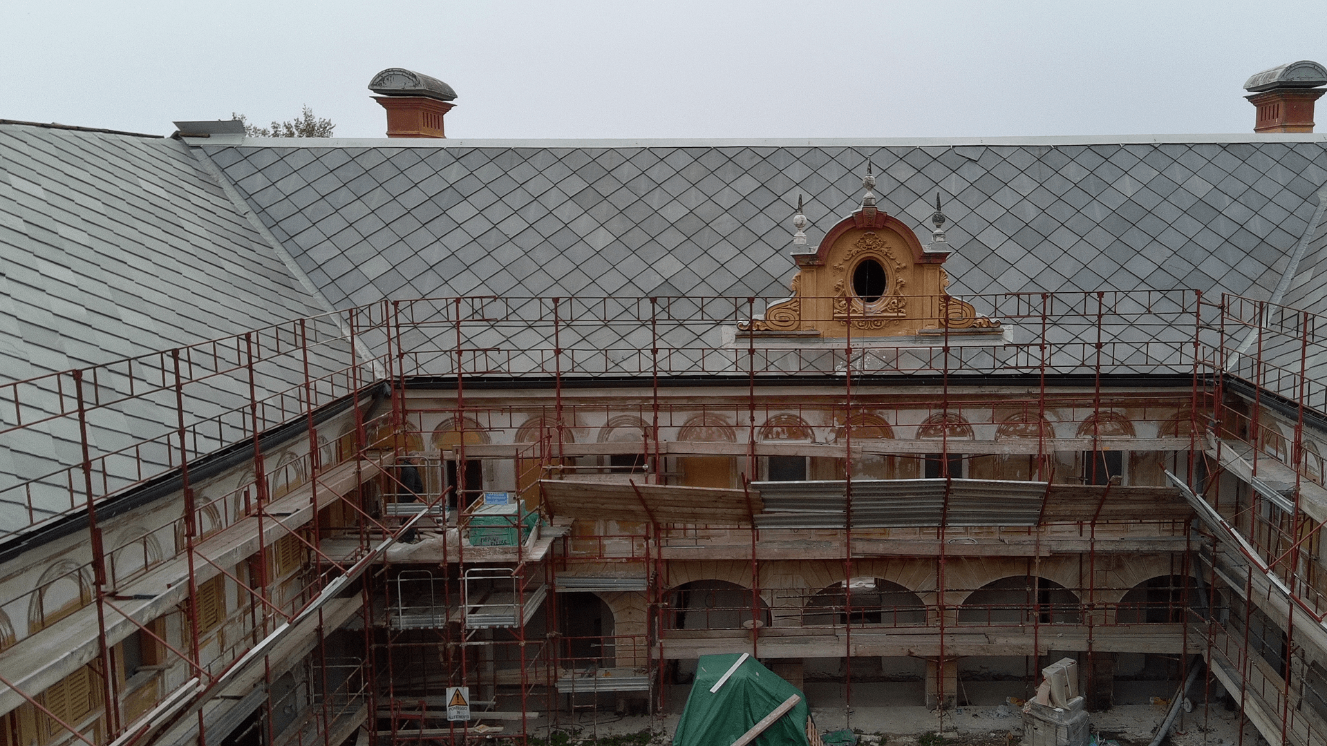 L'immagine mostra il tetto di un edificio storico in fase di ristrutturazione. Si può vedere una nuova copertura in tegole disposte in modo ordinato, che sta sostituendo o ripristinando quella esistente. L'edificio è parzialmente circondato da un'impalcatura rossa, che permette ai lavoratori di accedere alle zone più alte per completare il restauro e la ristrutturazione. In primo piano, possiamo notare una finestra decorata con dettagli architettonici e un elemento ornamentale sopra la finestra. La struttura stessa sembra essere in corso di restauro, con alcuni lavori di finitura sulla facciata inferiore visibile. Il tetto, in particolare, è stato quasi completato, mentre i lavori sulla facciata sembrano ancora in corso, con molte parti protette o smontate. Il contesto suggerisce che il restauro sta cercando di preservare il carattere storico dell'edificio, mantenendo gli elementi architettonici originali, come i dettagli intorno alle finestre e il tetto a forma di spiovente, mentre contemporaneamente viene eseguito il lavoro di modernizzazione.