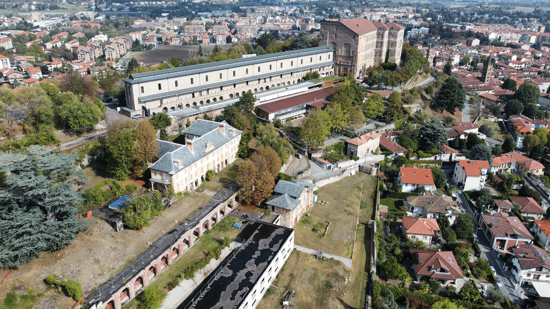 L'immagine mostra una vista aerea di un complesso architettonico che si trova su una collina, con edifici storici e una vista panoramica sulla città circostante. In primo piano, si vedono diversi edifici con tetti a falde rosse, situati in una zona residenziale. Al centro della scena, si trovano alcuni edifici storici, con il loro stile architettonico distintivo, con cortili verdi e una disposizione che suggerisce un ampio spazio. Il complesso più grande visibile è composto da un lungo edificio a più piani, situato su un terreno verde, con giardini e alberi che ne circondano i bordi. Accanto a questo edificio, si può notare una struttura con impalcature, probabilmente in fase di restauro, mentre dietro si erge un altro edificio più alto e massiccio con una facciata monumentale. Il panorama circostante è caratterizzato da una città a bassa densità edilizia, con case residenziali disposte lungo le colline. La vegetazione, gli spazi verdi e i sentieri che collegano il complesso storico con la zona residenziale indicano che il sito è stato progettato per integrarsi armoniosamente con il paesaggio. Questa scena offre una panoramica di come l'edificio storico e la zona circostante possano essere rinnovati o ripristinati, con l'aggiunta di modernità e comfort, senza perdere il legame con la sua storia e architettura originale.
